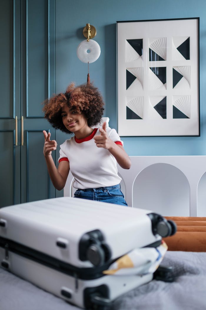 Curly-haired woman giving thumbs up beside packed luggage in a modern home.